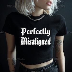 May include: Black crop top with the text "Perfectly Misaligned" in a gothic font. The shirt has short sleeves and a crew neck. The brand name "SIMPLYKEEAPPAREL" is printed on the top.