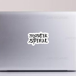 K&ouml;nnte beinhalten: Ein wei&szlig;er Laptop mit einem schwarzen Aufkleber auf dem Deckel, auf dem "MYSTIK SPIRAL" steht.