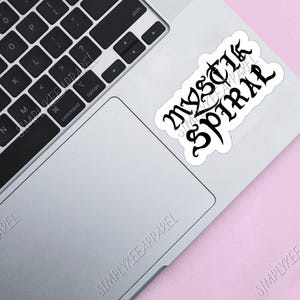 K&ouml;nnte beinhalten: Ein wei&szlig;er Laptop mit einer schwarzen Tastatur und einem schwarz-wei&szlig;en Aufkleber auf dem Trackpad, auf dem "MYSTIK SPIRAL" steht.