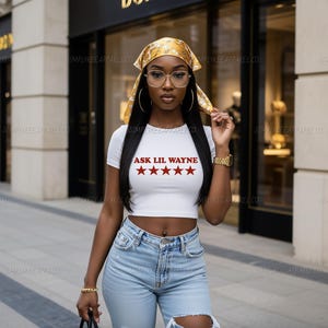 May include: A white short-sleeved crop top featuring the red text ASK LIL WAYNE above five red stars. The top is paired with light blue distressed denim jeans, a yellow patterned headscarf, gold hoop earrings, a gold watch, and gold bracelets.