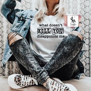 May include: A white t-shirt with the text "what doesn't kill you disappoints me" printed in black on it. The shirt is being worn by a person with black and white distressed jeans and a denim jacket.