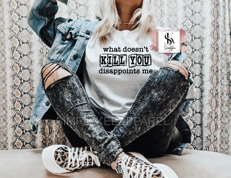 May include: A white t-shirt with the text "what doesn't kill you disappoints me" printed in black on it. The shirt is being worn by a person with black and white distressed jeans and a denim jacket.