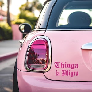 May include: Pink car with a black roof and chrome accents. The car has a sticker that says "Thinga la Migra" in a decorative font. The car's taillight is visible.
