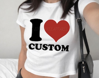 Op de afbeelding: Witte crop top met de tekst "I ♥ CUSTOM" in zwart en rood. De "I" is een zwarte pilaar, het hart is rood en het woord "CUSTOM" is in een vetgedrukte, zwarte letter. Een zwarte schoudertas hangt aan de schouder.