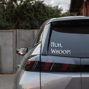 Puede incluir: Una calcomanía de vinilo blanco aplicada en la ventana trasera de un coche plateado. La calcomanía presenta el texto "Huh, WHOOP!" en una fuente sans-serif clara. El adhesivo está colocado en el lado derecho del cristal, contrastando con el tinte.