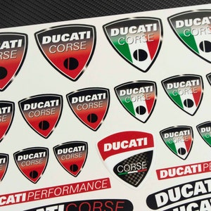 May include: A sheet of stickers featuring the Ducati Corse logo in various colors and designs. The stickers are printed on white paper and feature the Italian flag colors.