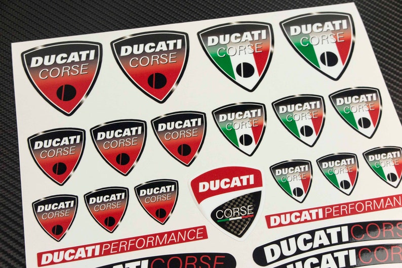 May include: A sheet of stickers featuring the Ducati Corse logo in various colors and designs. The stickers are printed on white paper and feature the Italian flag colors.