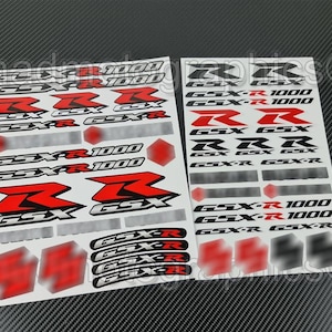 Puede incluir: Dos hojas de calcomanías para motocicletas con el texto "GSX-R 1000" y la letra "R" en rojo y blanco. Las calcomanías están diseñadas para una motocicleta y probablemente para personalización.