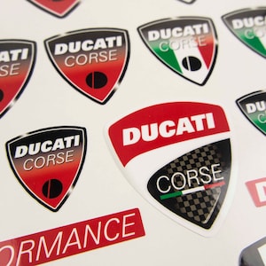 May include: A sheet of stickers featuring the Ducati Corse logo in various colors and designs. The stickers are printed on a white background and feature a red, white, and green Italian flag.