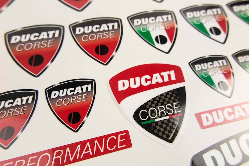 May include: A sheet of stickers featuring the Ducati Corse logo in various colors and designs. The stickers are printed on a white background and feature a red, white, and green Italian flag.