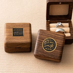 May include: Wooden ring boxes with personalized inscriptions. One box is open, revealing two rings. The other boxes have the names "Daniel & Sarah" and "Mr & Mrs Smith 05.23.26" engraved on them. The boxes are made of dark wood.