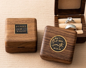 May include: Wooden ring boxes with engraved details. One box is open, displaying two rings. The other boxes feature the names "Daniel & Sarah" and "Mr & Mrs Smith 05.23.26". The boxes are made of dark wood with a warm tone.