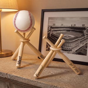 May include: Two wooden baseball bat stands, each crafted from three miniature bats joined by a ring. One stand holds a white baseball with red stitching. The bats are engraved with the names Albert and Emmanuel and the date Oct 8 2026.