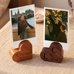 May include: Two heart-shaped wooden photo holders with slots for vertical prints. The left holder is light brown wood engraved with "Crystal" and "08.06.2026". The right holder is dark brown wood engraved with "L & C" and "08.11.26".