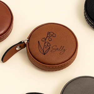 Könnte beinhalten: Ein braunes, rundes Maßband mit einem schwarzen Blumendesign und dem Namen "Sally" in Schreibschrift. Das Maßband hat eine Lederlasche und einen Metallring. Weitere braune und schwarze Maßbänder sind im Hintergrund.