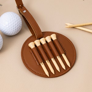May include: A brown leather golf tee holder with a loop for attaching to a golf bag. The holder has six slots for golf tees, each with the word "SON" engraved on the tee.