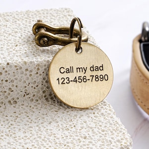 May include: A round, gold-colored pet tag with the engraved text "Call my dad 123-456-7890". The tag is attached to a bronze-colored clasp. The background is a textured, off-white surface.