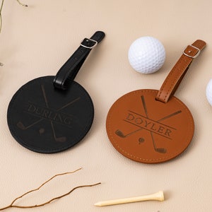 May include: Two round leather luggage tags with black and brown straps. The black tag has the name "DURLING" engraved on it and the brown tag has the name "DOYLER" engraved on it. Both tags have a design of two crossed golf clubs.