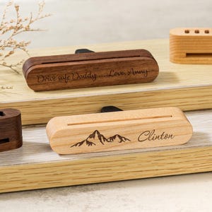 Könnte beinhalten: Handyhalter aus Holz mit eingravierten Designs. Einer ist dunkelbraun mit dem Text "Drive safe Daddy... Love, Anny". Ein anderer ist hellbraun mit einem Bergmotiv und dem Namen "Clinton".