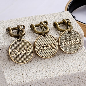 May include: Three bronze-colored pet ID tags with different engraved names: "Buddy", "Berry", and "Nova". Each tag is round with a decorative pattern and attached to a bronze clasp. The tags are displayed on a textured, light-colored surface.