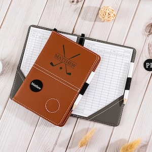 May include: A brown leather golf score card holder with the name "MATTHEW" and golf club graphics. The scorecard holder is open, revealing a scorecard and a pencil. The holder is on a wooden surface.