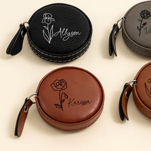May include: Several personalised round tape measures in black, grey, and brown. Each has a floral design and a name, such as "Allyson" and "Karissa", in cursive script. The tape measures have a leather-like texture and a white stitched border, with a small leather strap.