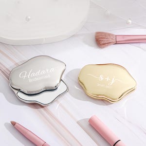 May include: Two compact mirrors with irregular edges. One is silver and engraved with "Hadara Bridesmaid." The other is gold, inscribed with "s + j" and the date "20.07.2027." Makeup brushes and tools are in the background. Wedding favors.
