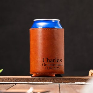 May include: A brown leather can cooler with the name "Charles" and the text "Groomsman 11.08.2025" engraved on it.