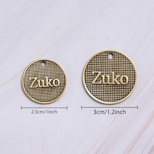 May include: Two gold-colored round charms with the name "Zuko" embossed on them. The charms have a textured grid pattern and a small hole at the top. One charm is 1 inch in diameter, and the other is 1.2 inches.