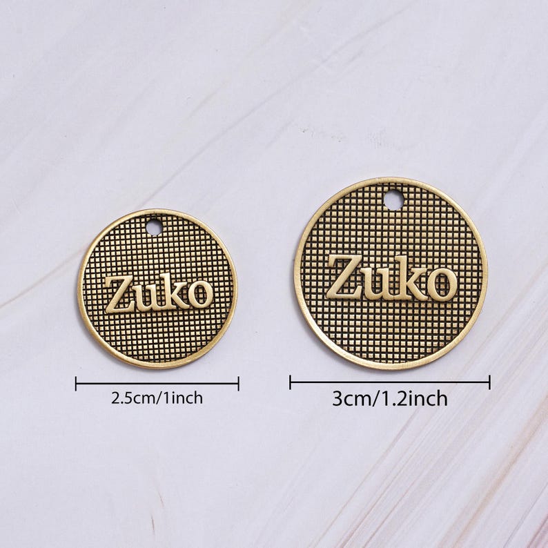 May include: Two gold-colored round charms with the name "Zuko" embossed on them. The charms have a textured grid pattern and a small hole at the top. One charm is 1 inch in diameter, and the other is 1.2 inches.