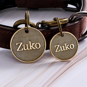 May include: Two round, bronze-colored dog tags with the name "Zuko" in a bold font. The tags have a textured grid pattern and are attached to a brown leather dog collar with bronze hardware.