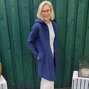 Könnte beinhalten: Eine Frau in einem blauen Kapuzenmantel, einer weißen Hose und weißen Turnschuhen steht vor einem grünen Holzzaun. Sie lächelt und schaut in die Kamera.