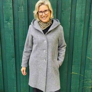 Könnte beinhalten: Ein grauer Mantel mit Kapuze und drei Knöpfen. Der Mantel hat eine taillierte Passform und Taschen. Die Person trägt einen Leoparden-Schal und eine schwarze Hose. Der Hintergrund ist ein grüner Holzzaun.