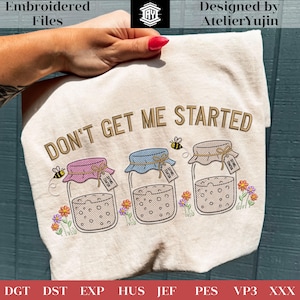 May include: A beige t-shirt with the embroidered phrase "DON'T GET ME STARTED" above three jars with colorful lids and tags. The jars are decorated with bees and flowers. The design is by AtelierYujin.