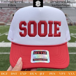 May include: A white and red trucker hat featuring the word SOOIE embroidered in red block letters on the front panel. The hat has a white mesh back, a red brim with a decorative cord, and a holographic sticker on the brim reading OTTO CLASSIC.
