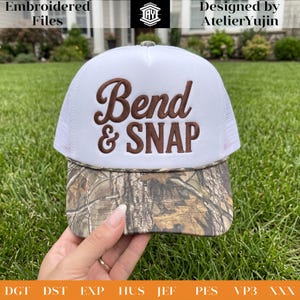 May include: A white trucker hat featuring a brown embroidered text that reads Bend & SNAP on the front foam panel. The hat has a camouflage patterned brim with a thin brown cord detail separating the brim from the white mesh back panels.