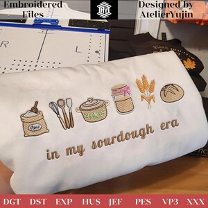 Può includere: Tessuto bianco ricamato con illustrazioni a tema panetteria e la scritta "in my sourdough era". Il design include un sacco di farina, frusta, pentola, barattolo, grano e pane. Design di AtelierYujin.