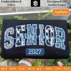 May include: Black sweatshirt with the words "SENIOR mom 2027" embroidered in blue and leopard print. The design includes the text "Embroidered Files" and "Designed by AtelierYujin". The word "Applique" is also visible.