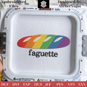 Op de afbeelding: Een witte stof in een borduurring toont een regenboogkleurig baguette-ontwerp met het woord "faguette" eronder geborduurd. De teksten "Embroidered Files" en "Designed by AtelierYujin" zijn zichtbaar. Gemaakt in de VS.