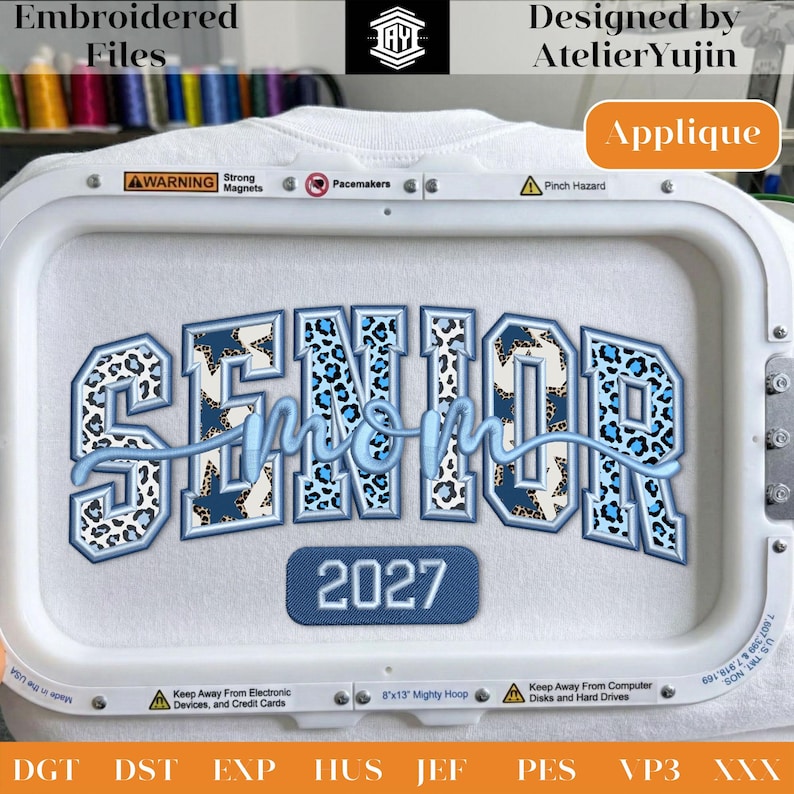 May include: White t-shirt with an embroidered design. The design features the words "SENIOR MOM" in blue and leopard print, with the year "2027" below. The design is set within an embroidery hoop.
