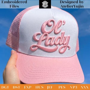May include: A pink and white trucker hat with the embroidered text "Ol' Lady" in a cursive font. The hat has a pink brim and a white front panel, with a mesh back. The words "Embroidered Files" and "Designed by AtelierYujin" are also visible.