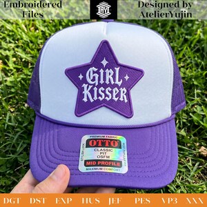 May include: A purple and white trucker cap featuring a purple star-shaped patch on the front panel with the white embroidered text Girl Kisser. The cap has a purple brim, a white foam front, and a purple mesh back with a sticker on the brim.
