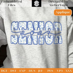 Può includere: Una felpa grigio chiaro con le parole "SENIOR 2027" ricamate in blu con un motivo floreale. Il design include il testo "Embroidered Files" e "Designed by AtelierYujin". È visibile anche un'etichetta "Applique".