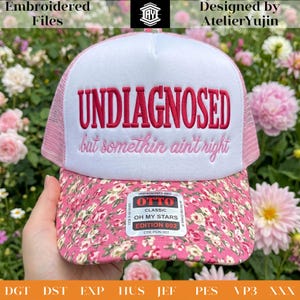 Può includere: Un cappellino da camionista bianco e rosa con la scritta ricamata "UNDIAGNOSED but somethin ain't right". Il cappello presenta un bordo a fantasia floreale e un retro in rete rosa. Il cappello è un "CLASSIC OH MY STARS EDITION 002" di OTTO.