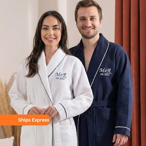 May include: Two waffle-weave bathrobes, one white and one navy blue, with dark blue trim. Each robe has the embroidered initials "M&R" and the year "est.2022". The white robe is worn by a person on the left, and the navy robe is worn by a person on the right.