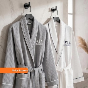 May include: Two waffle-weave bathrobes, one gray and one white, hang on black hooks. Each robe has a matching belt, pockets, and white trim. The gray robe has white trim, and the white robe has navy trim. Both robes are embroidered with "K|L Est.2026".