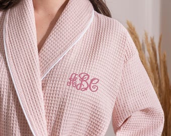 May include: A light pink waffle-weave robe with white trim and a monogram in a matching pink thread. The robe is made of a textured fabric and has a shawl collar. The monogram features the letters "ABC" in a cursive font.