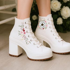 May include: White high-top lace-up boots with a block heel and platform sole. The boots feature delicate floral embroidery in shades of pink, red, and green on the side, and a circular logo patch on the ankle with a star and floral wreath.