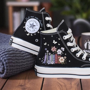 May include: Black high-top sneakers with white laces and soles. The sneakers feature floral and book designs, along with the text "Just one more chapter." A coffee mug and a folded gray blanket are in the background.