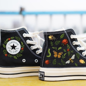 Könnte beinhalten: Schwarze High-Top-Sneaker mit weißen Schnürsenkeln und Sohlen. Die Sneaker zeigen detaillierte Illustrationen von Insekten und Blättern. Ein Schuh hat ein Kranzdesign mit dem Text "Converse All Star".